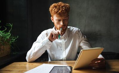 Photo of young thinking readhead bearded man, holding notebook and green pan, sitting at wooden table